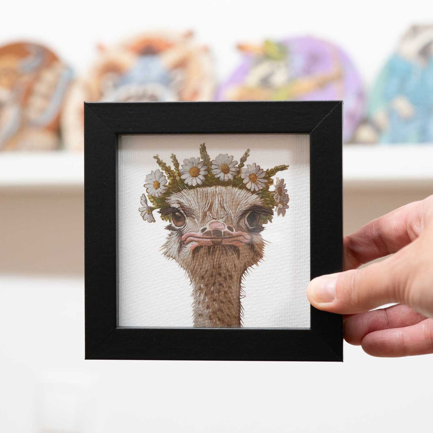 Hand holding a framed fine art print of a flower-crowned ostrich, perfect for brightening up small spaces.