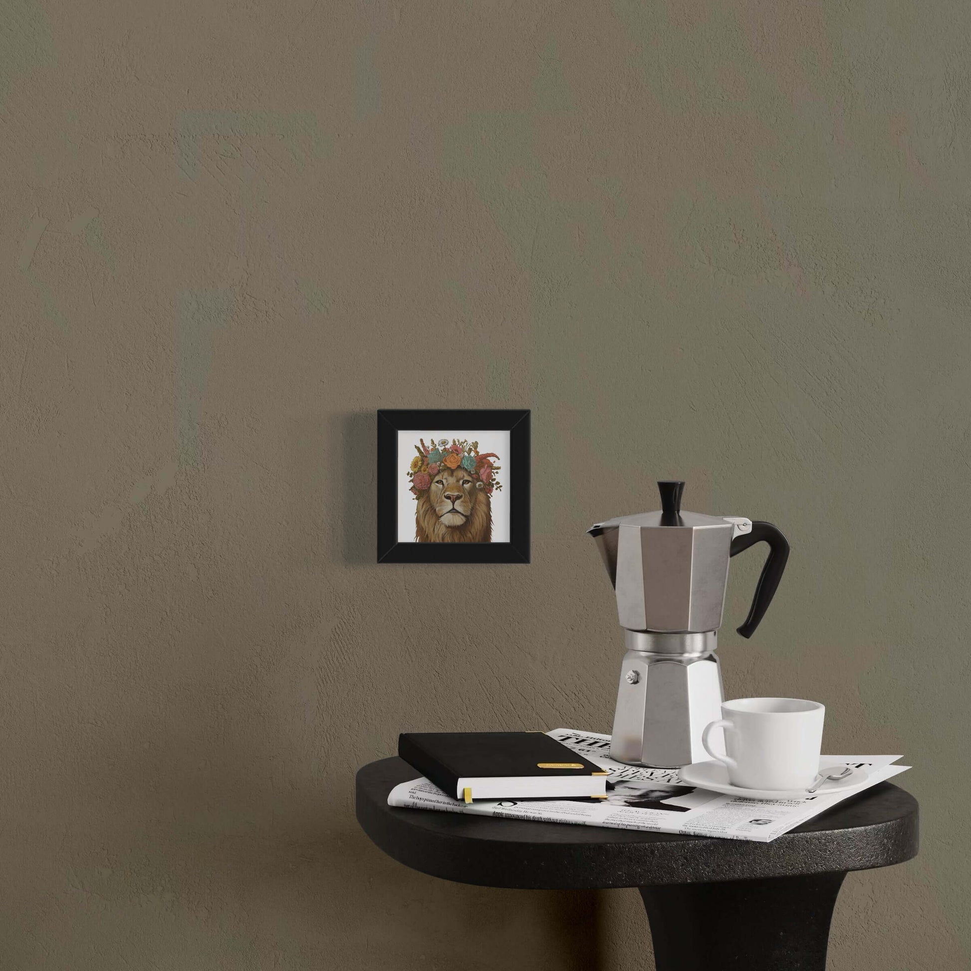 Fine art print of a lion with floral crown, framed on a wall beside a coffee pot and notebook.