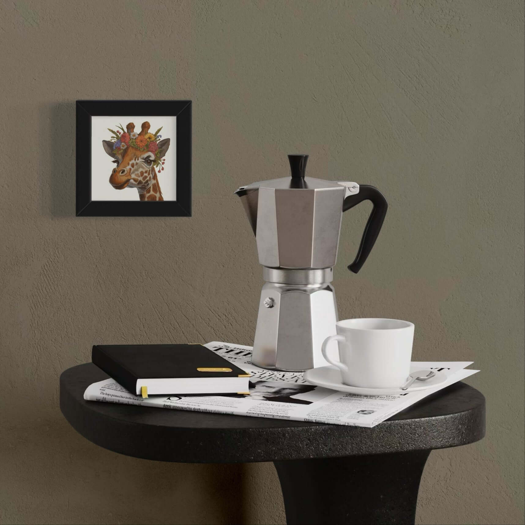 Framed giraffe art print with floral crown on wall above coffee maker and notebook on black table.