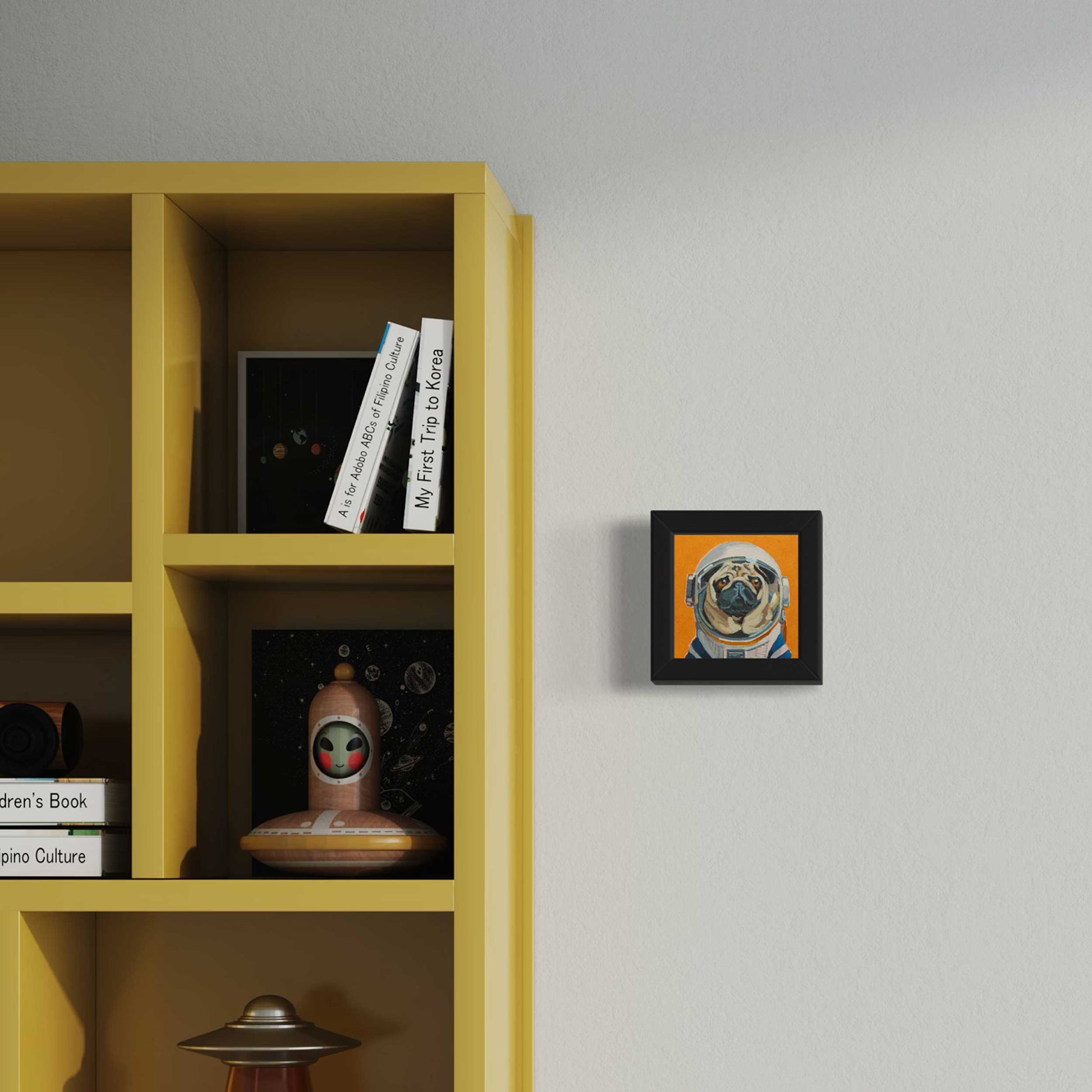 Gerahmter Fine Art Print Astro Mops 10x10 cm, auf Hahnemühle Büttenpapier neben gelbem Bücherregal.